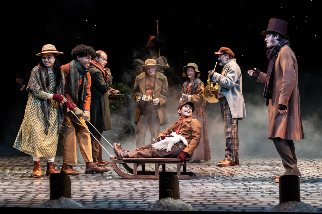 A boy on a sled is center stage, surrounded by a cast of actors in period costumes